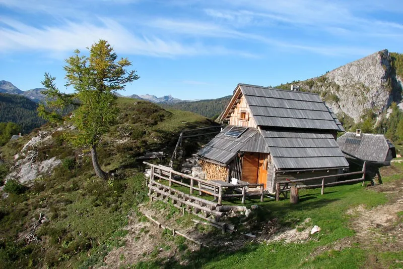 Lesena koča s skodlasto streho na planini Krstenica, za njo skalnat greben